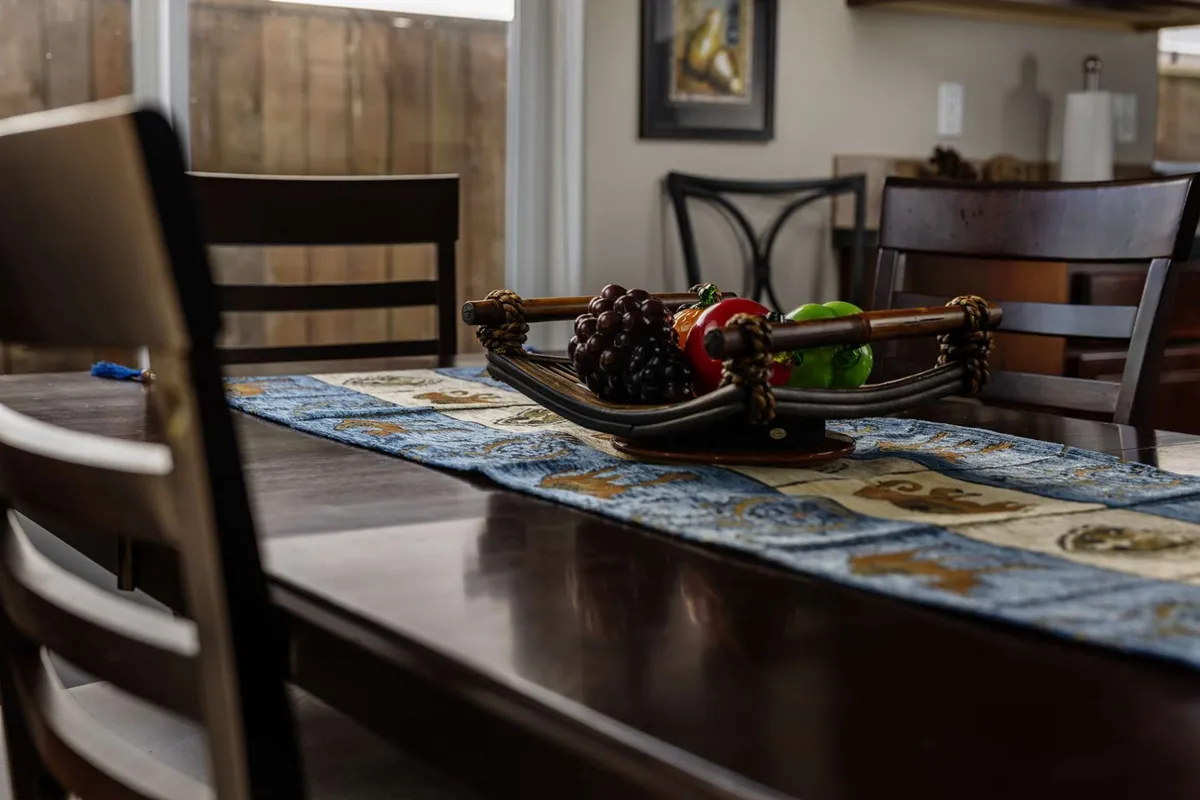 A welcoming dining room scene featuring a well-set table with a fruit basket, a decorative table runner, and surrounding chairs. The space radiates warmth and is clearly designed for gatherings, meals, and celebrations.
