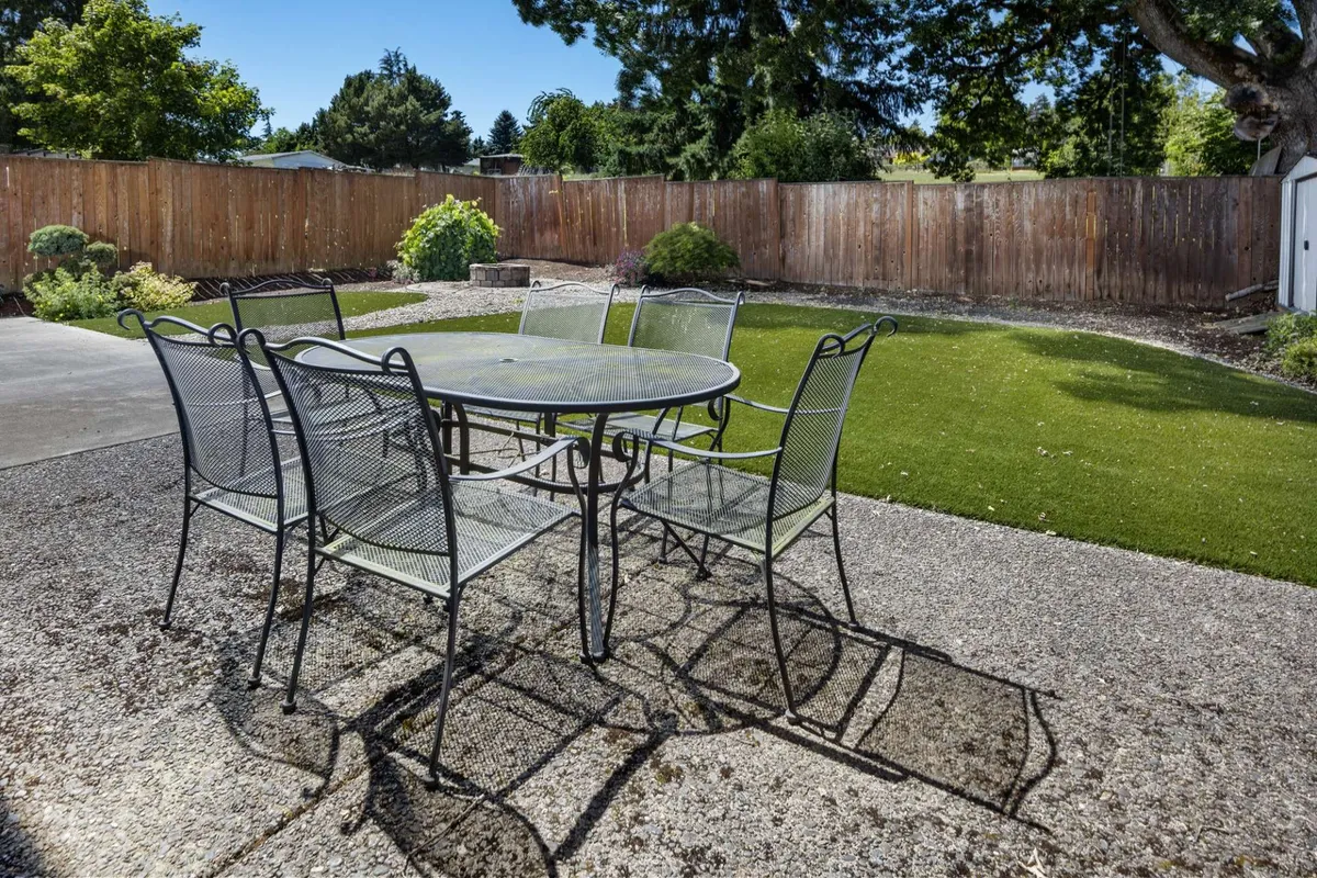 This image showcases a welcoming backyard scene featuring an outdoor dining table set on a patio, surrounded by lush greenery and enclosed by a fence. The area is clearly designed for alfresco meals, family gatherings, and relaxation in a sunny private environment. The fenced perimeter and well-maintained lawn offer both security and space for play or leisure.