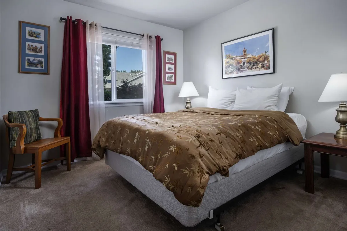 A peaceful bedroom scene featuring a bed with brown bedding, a window bathing the room in natural light, decorative wall art, a side table with a lamp, and an armchair. The space looks serene and ideal for relaxation or enjoying a quiet morning coffee.