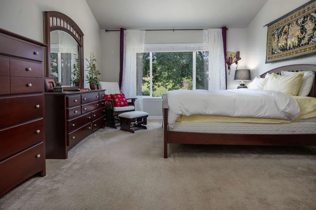This image showcases a serene bedroom with a bed adorned in yellow accents, a large window letting in natural light, a wooden dresser, and a decorative wall hanging. The setting is tranquil and inviting, capturing the essence of a restful retreat.